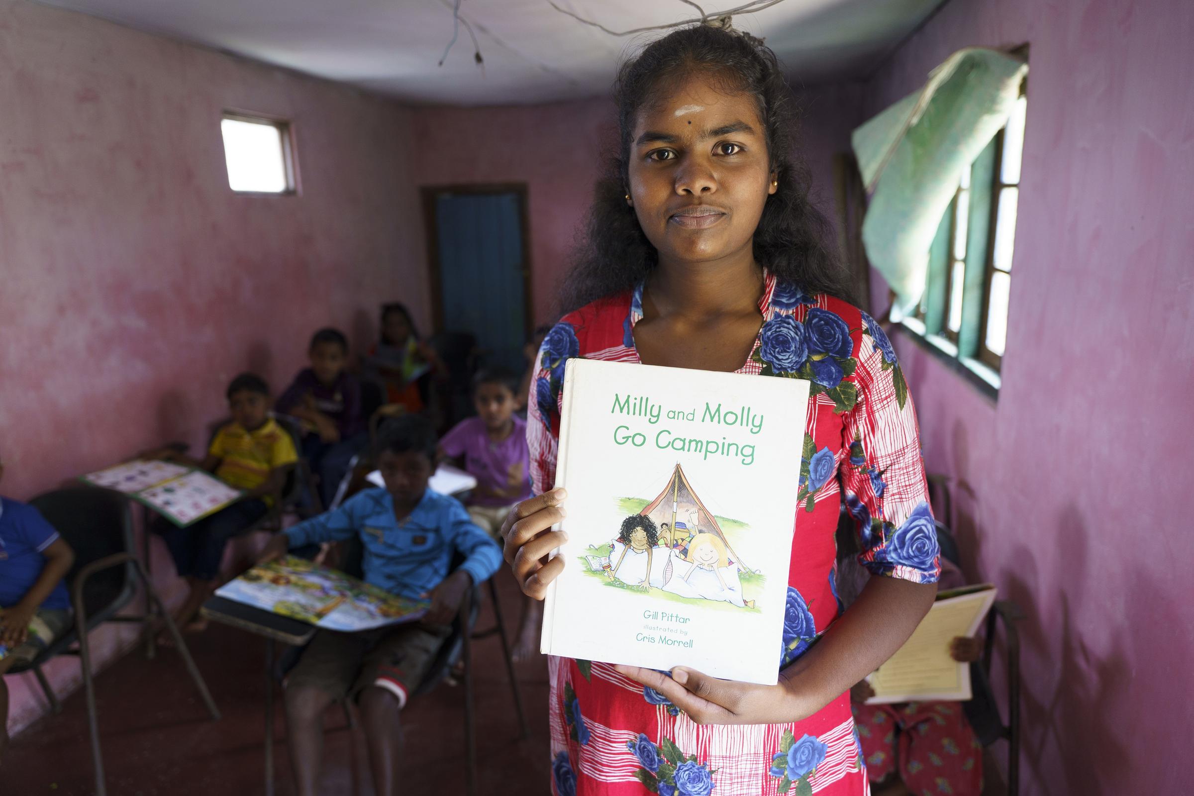 Eine Junge Frau steht in einer Bücherei und hält ein Buch vor sich hoch (Quelle: Christian Nusch) Die Lehrerin hilft, wenn ein Kind etwas nicht versteht (Foto: Christian Nusch)
