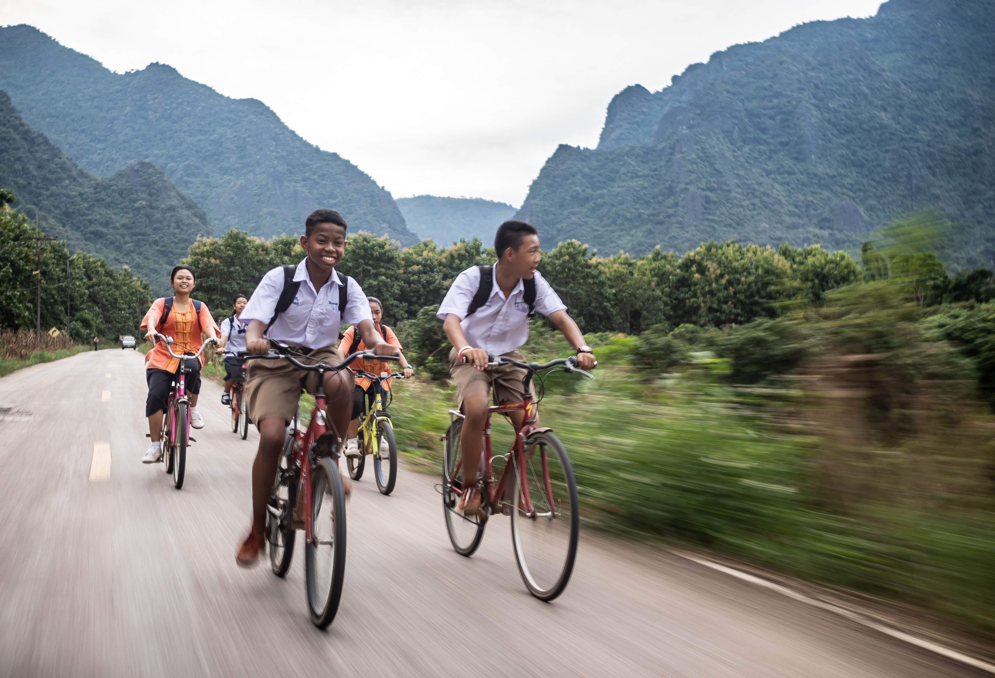 Eine Gruppe jugendlicher Radfahrerinnen und Radfahrer fährt zügig eine Straße entlang, im Hintergrund Dschungel und hohe Berge  (Quelle: Jakob Studnar)