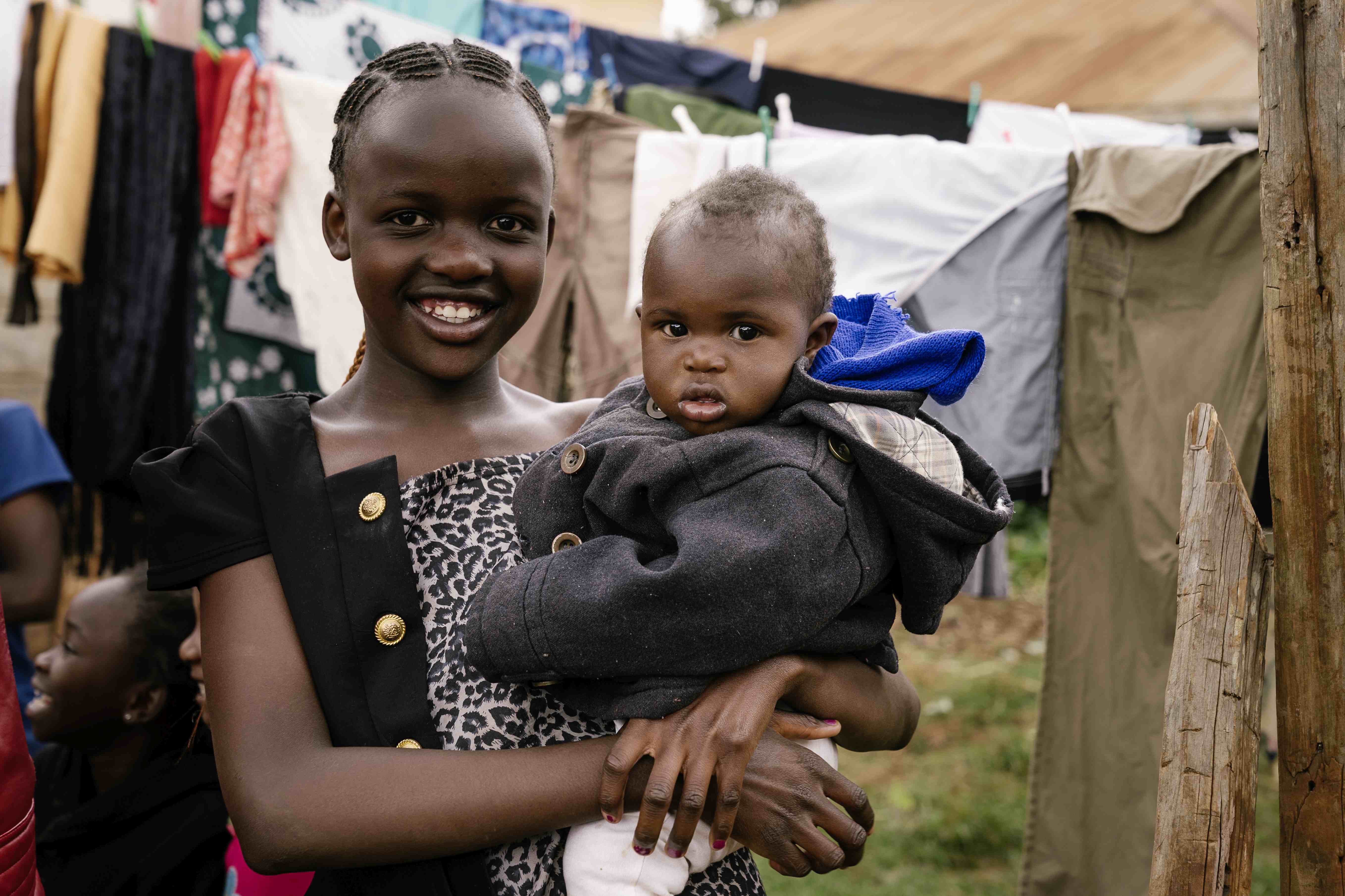 Mädchen mit Baby auf dem Arm steht draußen vor einer Wäscheleine mit Wäsche  (Bildquelle: Dan Zoubek)